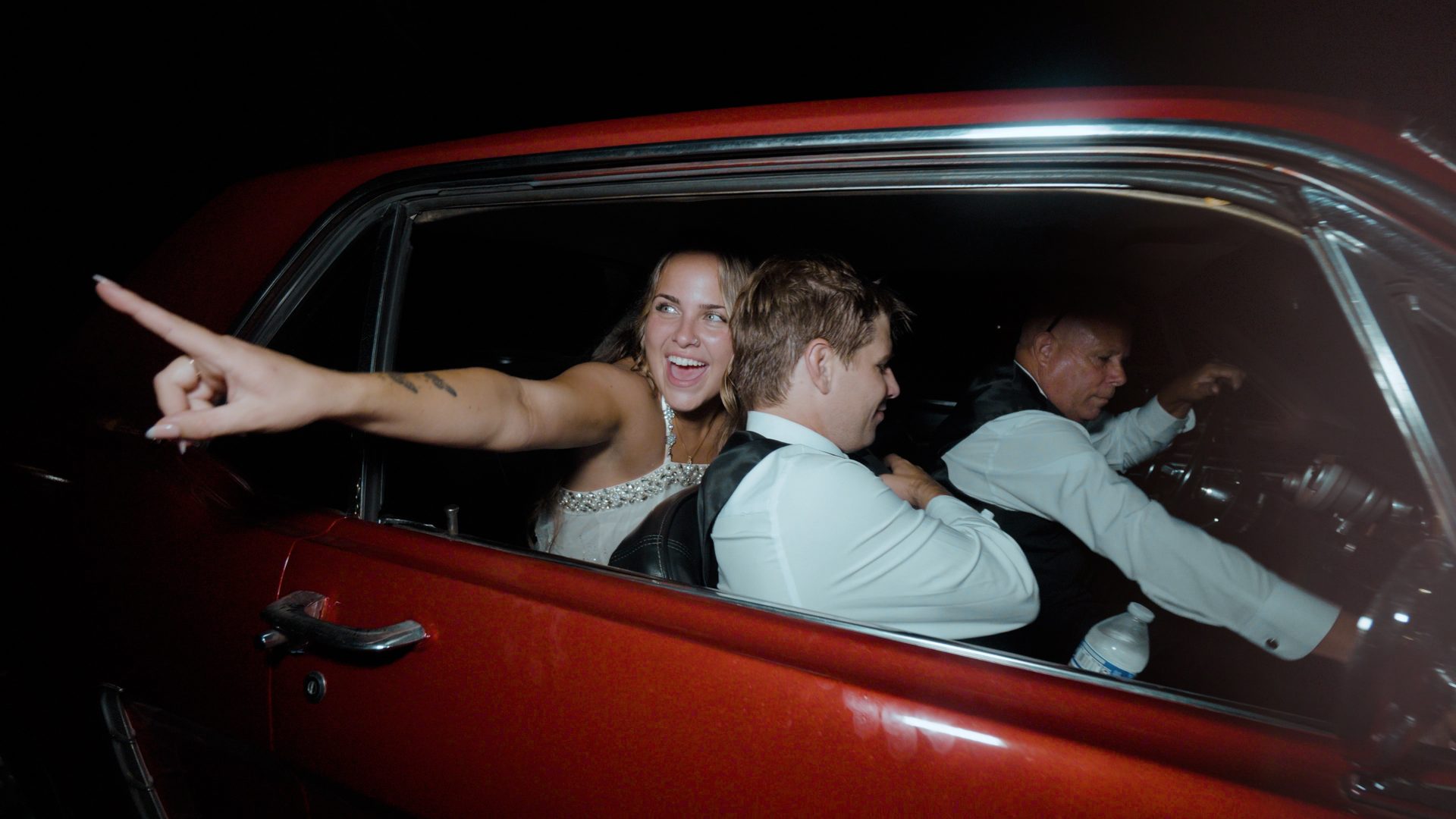 Bride laughing and pointing from a red vintage car during a wedding send-off